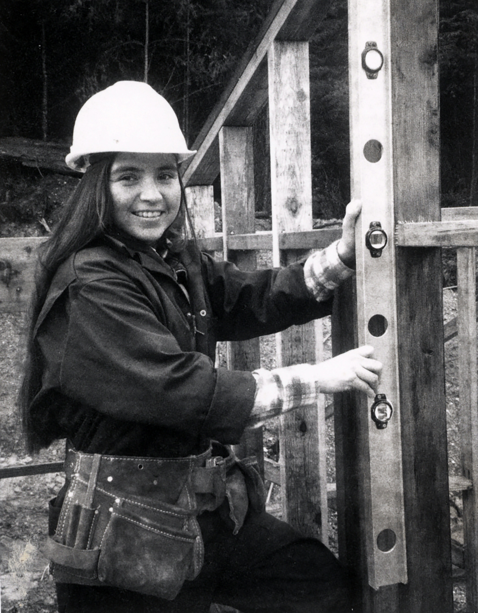 Toni Whipple wears a hard hat and tool belt as she holds a level up to a timber framing.