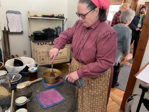A woman stands in a kitchen in a period dress. She stirs thick maple syrup in a pot.