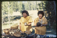 Two women stand at a table holding woven baskets in front of a meadow. 
