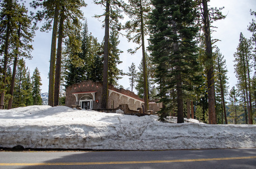 A stone building surrounded by snow and tall conifer trees. A road passes across the foreground.