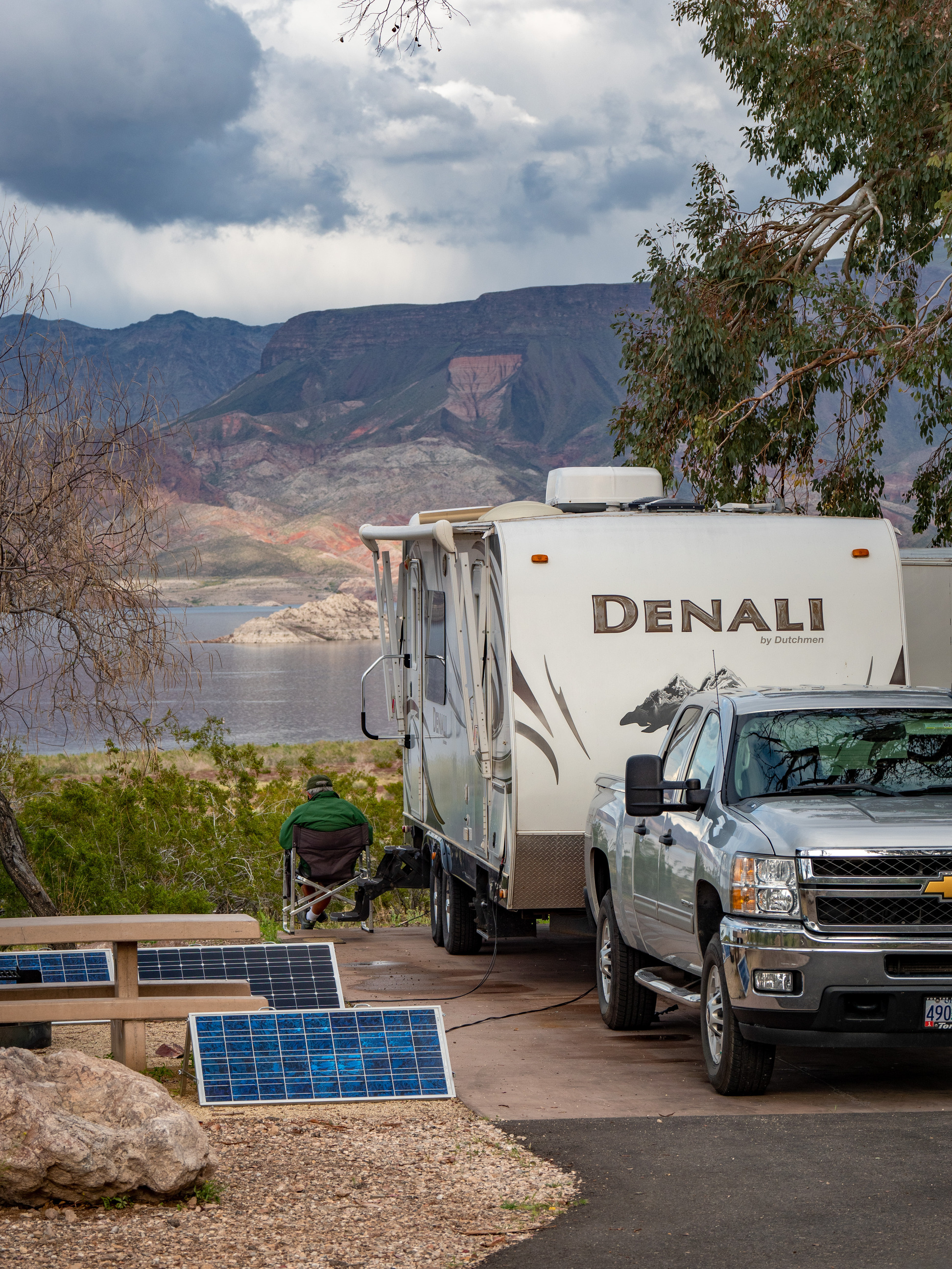 truck and camp trailer from front at right, person in chair and small solar panels at left, lake and mountains in distance