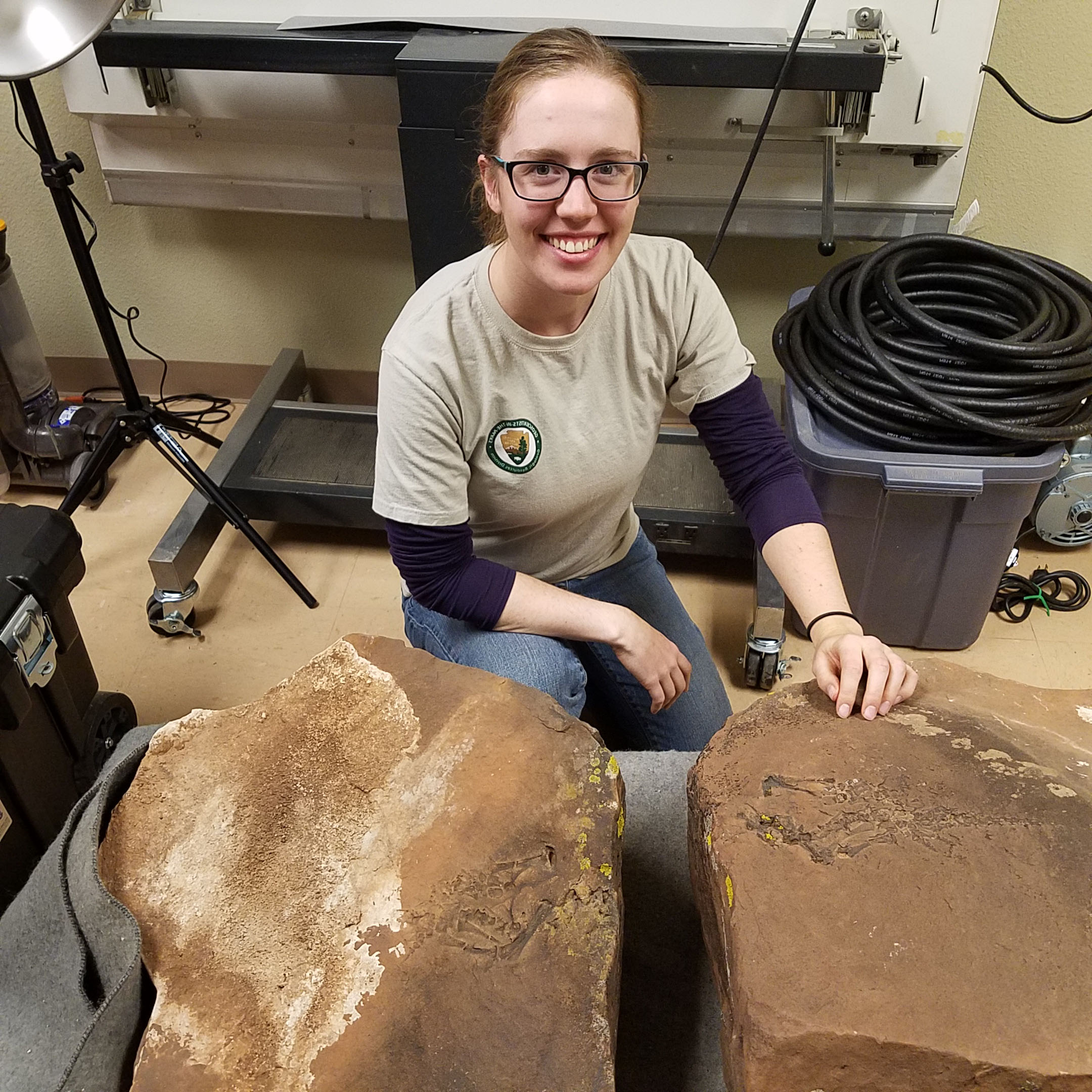 A young woman with glasses kneels next to two large fossils in a laboratory. 