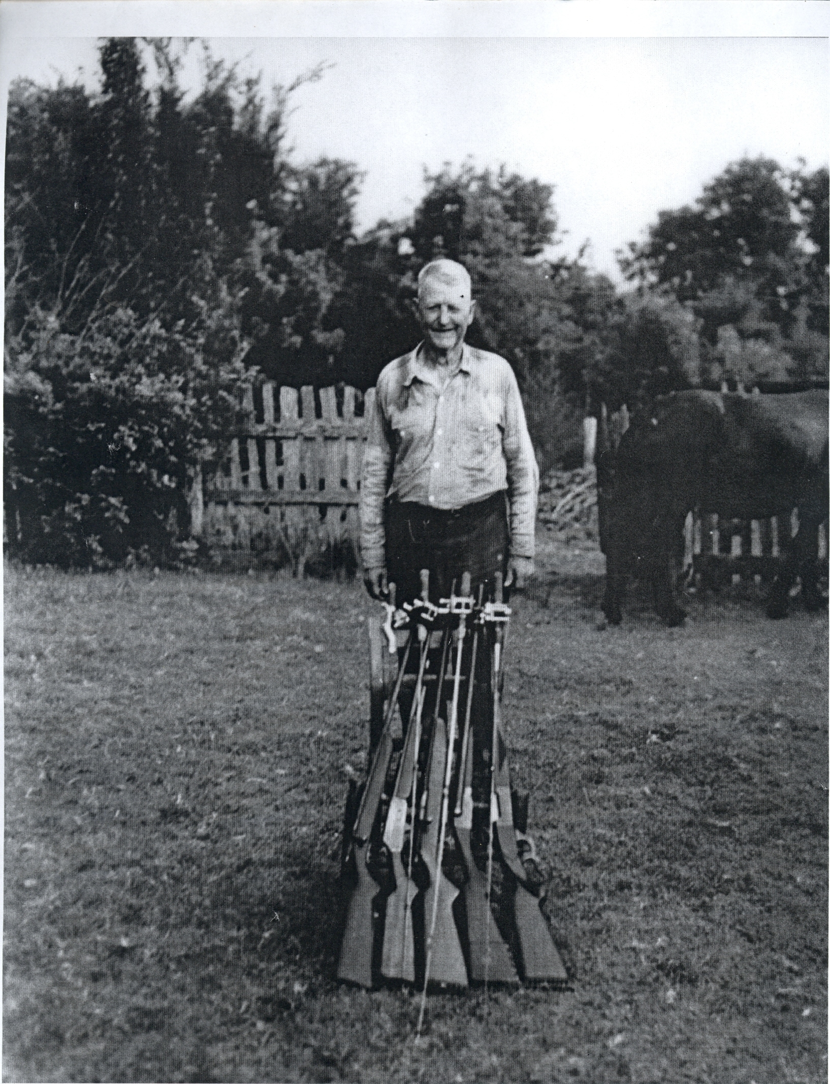 black and white photo of man in yard with rifles and fishing poles in front of him, back half of horse and grey picket fence behind him