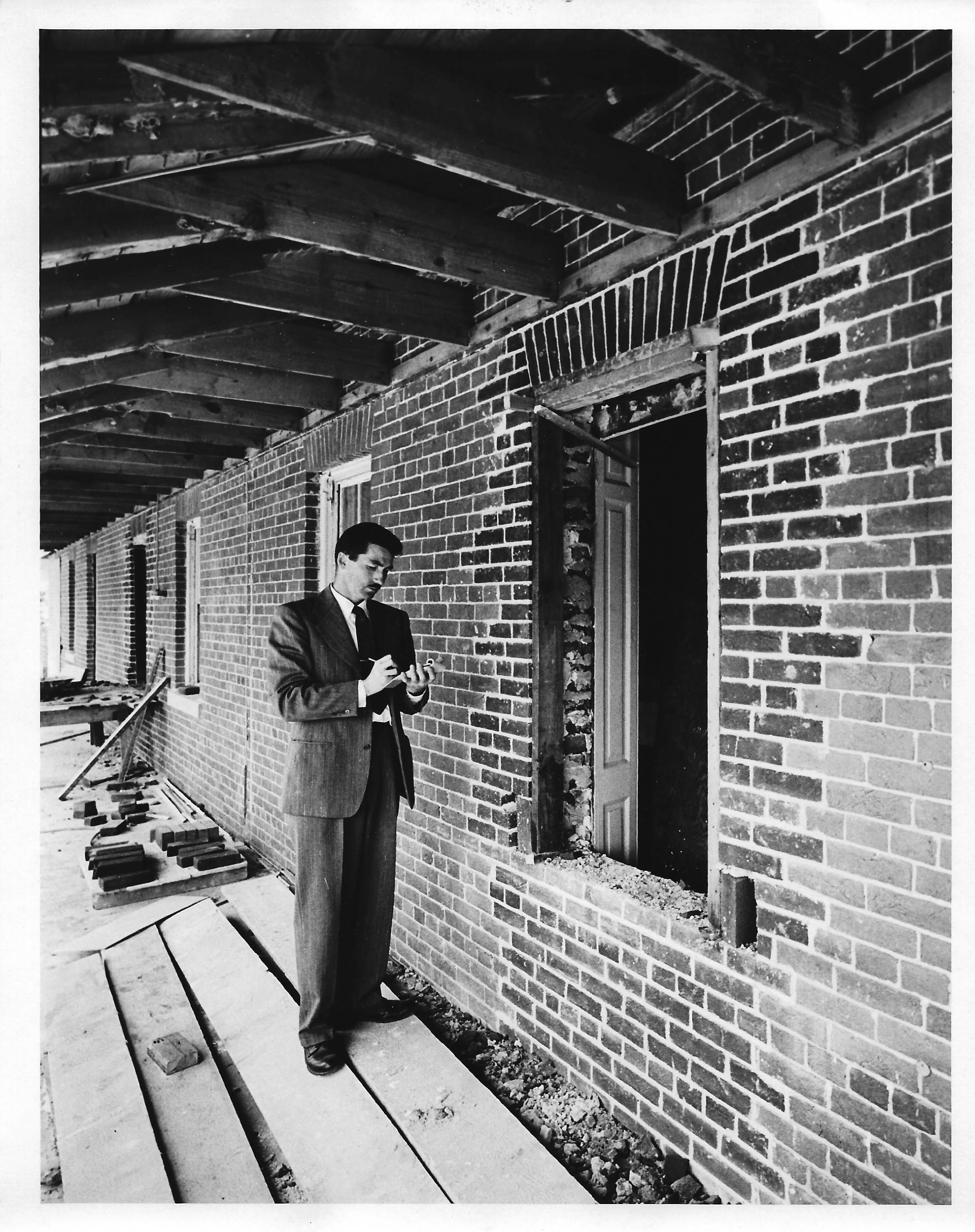 Man in suit inspecting the brickwork that was done to fill in the doorway.  .  Standing  on a wood path that was built, behind him are bricks and tools on the path or leaning against the wall.