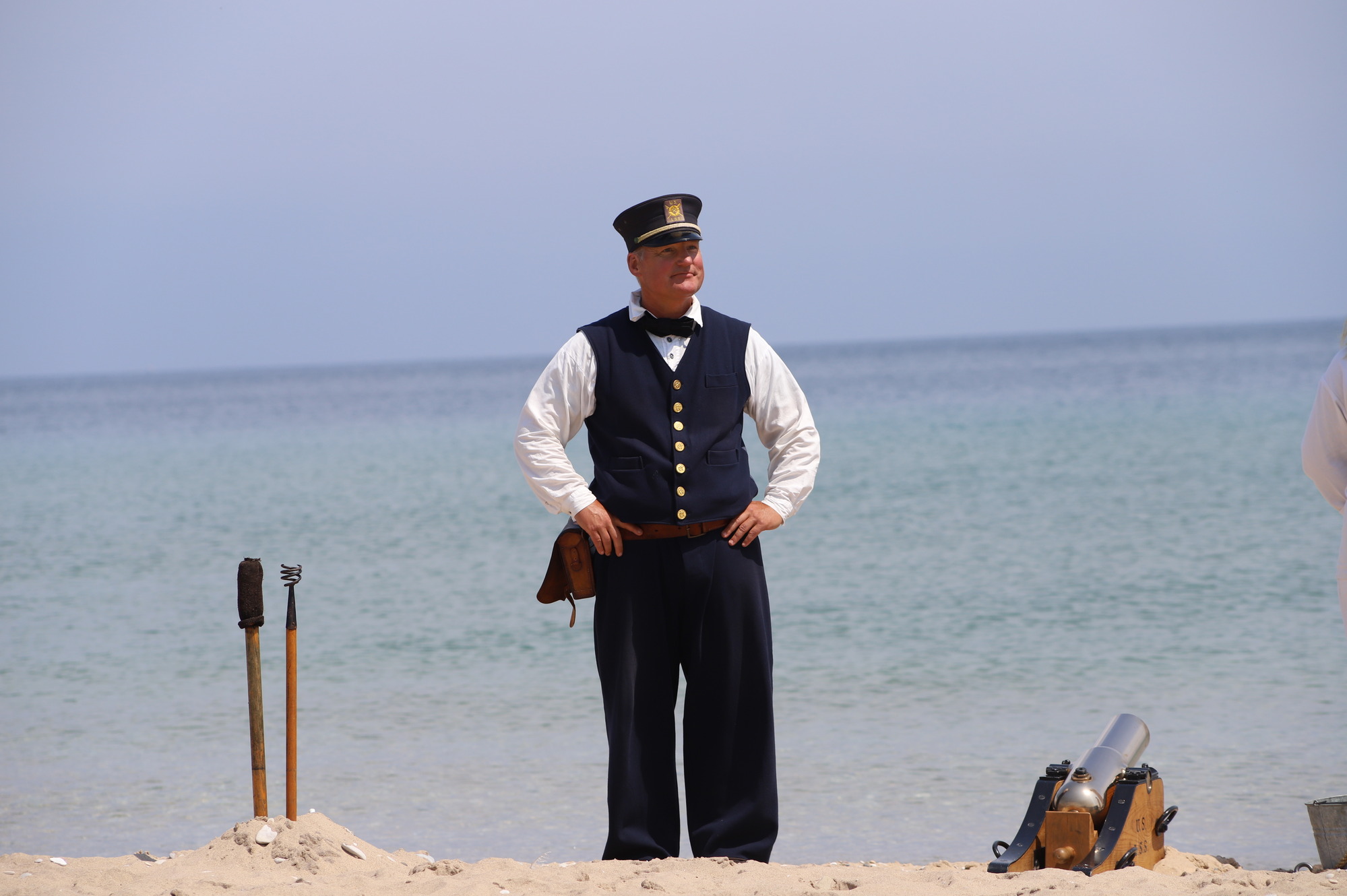 Keeper Matt in his navy-blue uniform stands heroically with his trusty, lifesaving Lyle Gun to one side and tools to the other. The blue waters of Lake Michigan stretch to the horizon behind him.