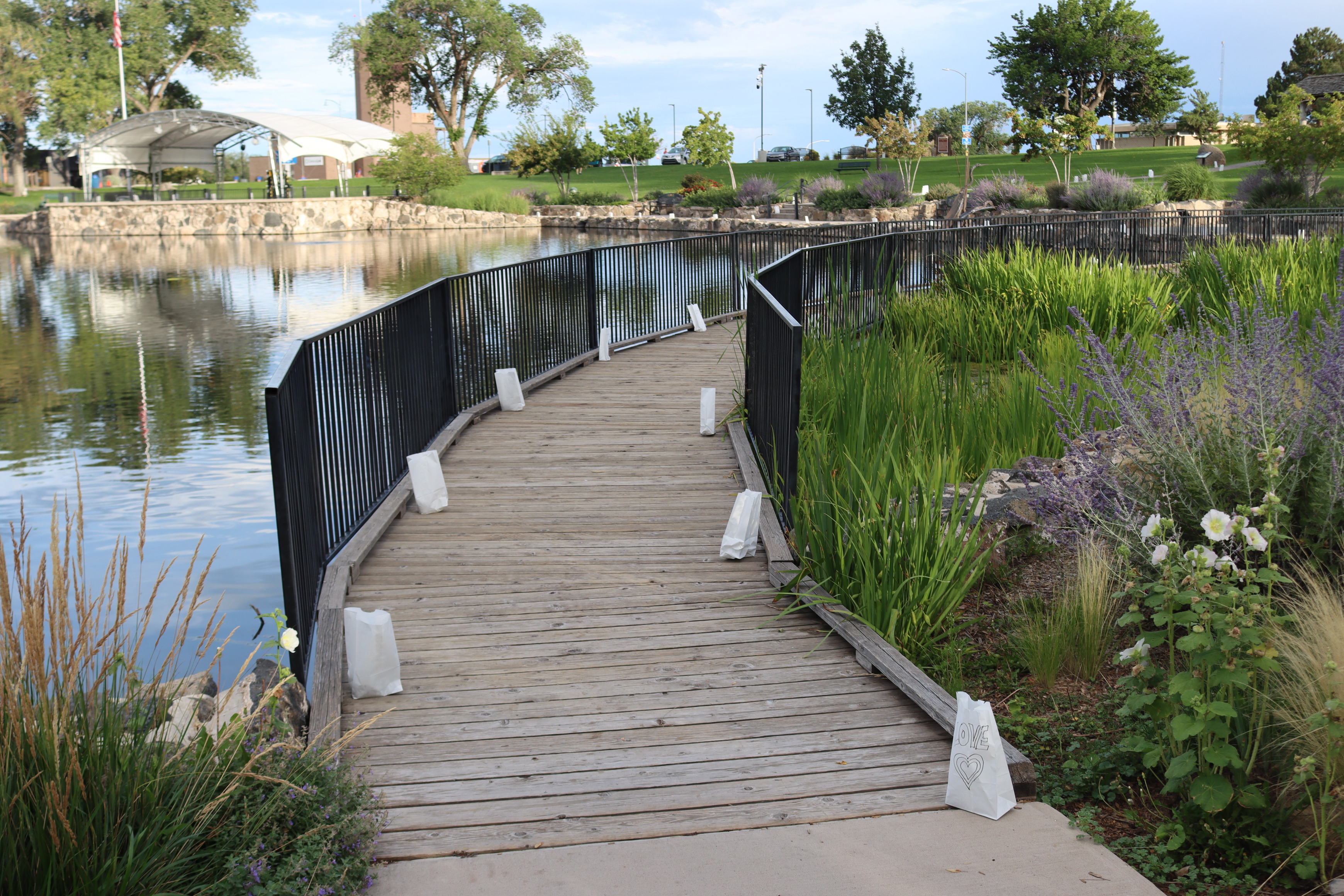 On a reflective pond, a wooden boardwalk and black metal railings trails around green blooming pond plants. A white covered stage and manicured park are across the water in the background.