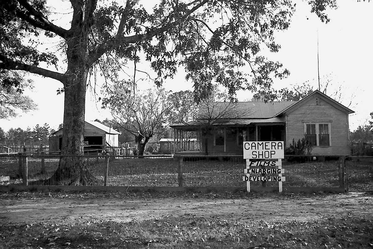Black and white photo of a sign outside a house next to a big tree. The sign reads: Camera Shop; films, enlarging, developing.
