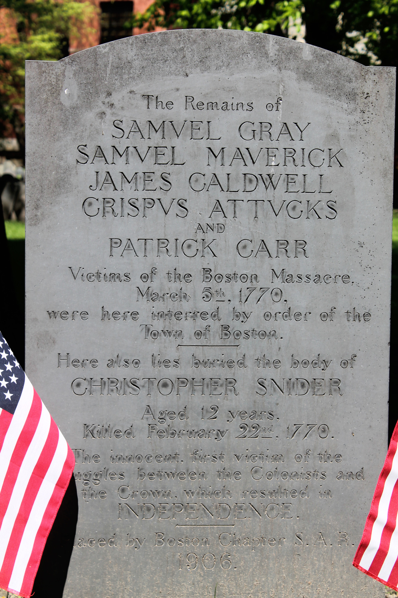A close up of the grave of the victims of the Boston Massacre and Christopher Snider or Seider. The marker is grey in color and has little texture to it. The text carved on it is: The Remains of Samuel Gray, Samuel Maverick, James Caldwell, Crispus Attucks, and Patrick Carr...Victims of the Boston Massacre, March 5th, 1770, were here interred by order of the Town of Boston. Here also lies buries the body of Christopher Snider Aged 12 years killed February 22, 1770. The innocent first victim of the struggles between the Colonists and teh Crown, which resulted in Independence. Placed by Boston Chapter S.A.R. 1906.
On either side of the bottom of the marker are small American flags.