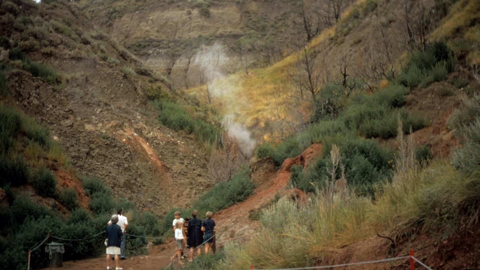 Five people on a trail look towards smoke rising from the ground in a steep gully.