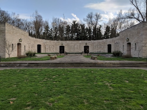 Limestone block building, half circle shape with six openings spaced evenly apart. Light fixtures above four openings. Two windows on each side of left opening and right opening. Green grass in foreground leads to stairs and walkway with stone benches on each side. Bare trees and evergreens in background. Blue sky with clouds.