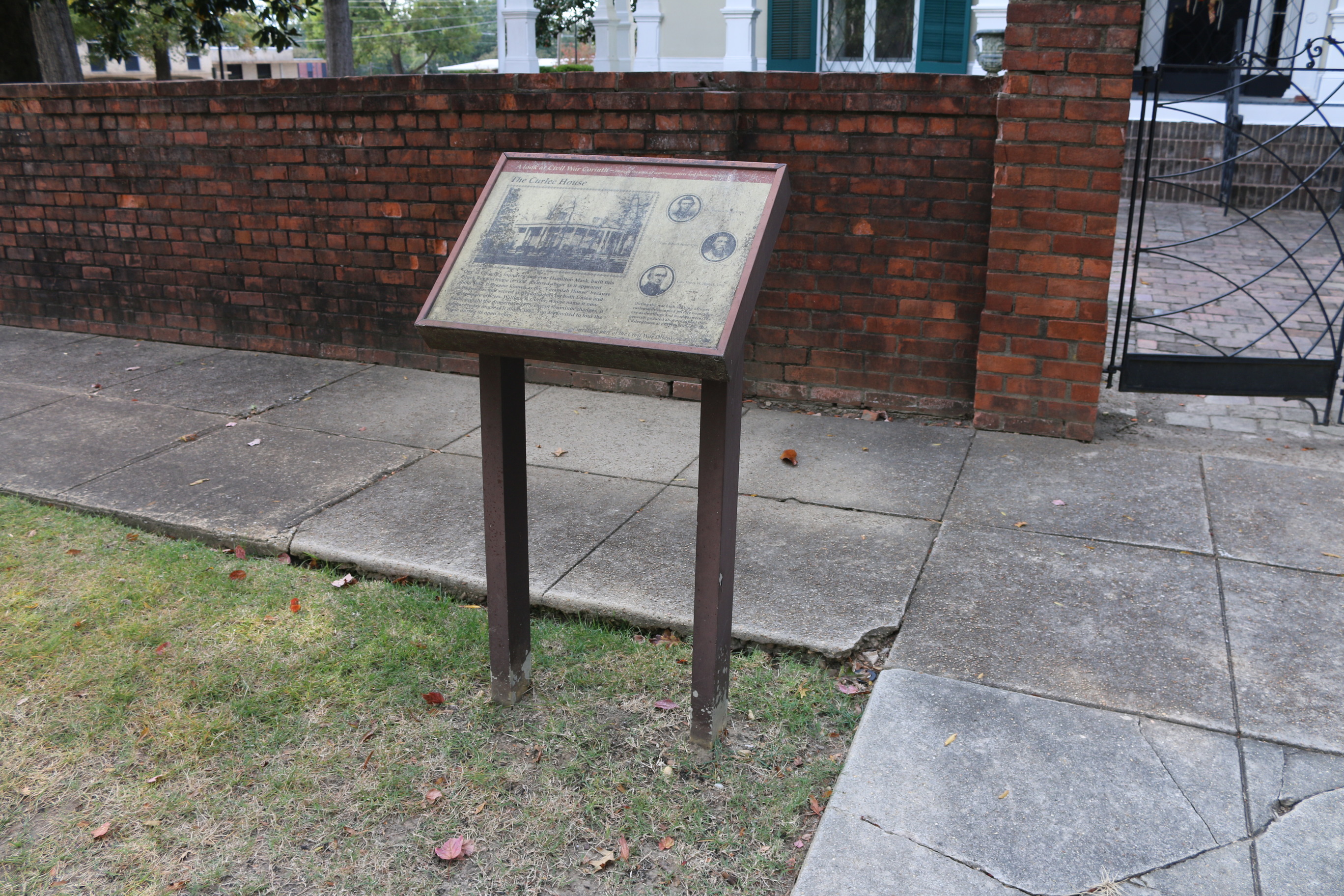 A battered wayside stands alongside a sidewalk and brick wall before a historic home.