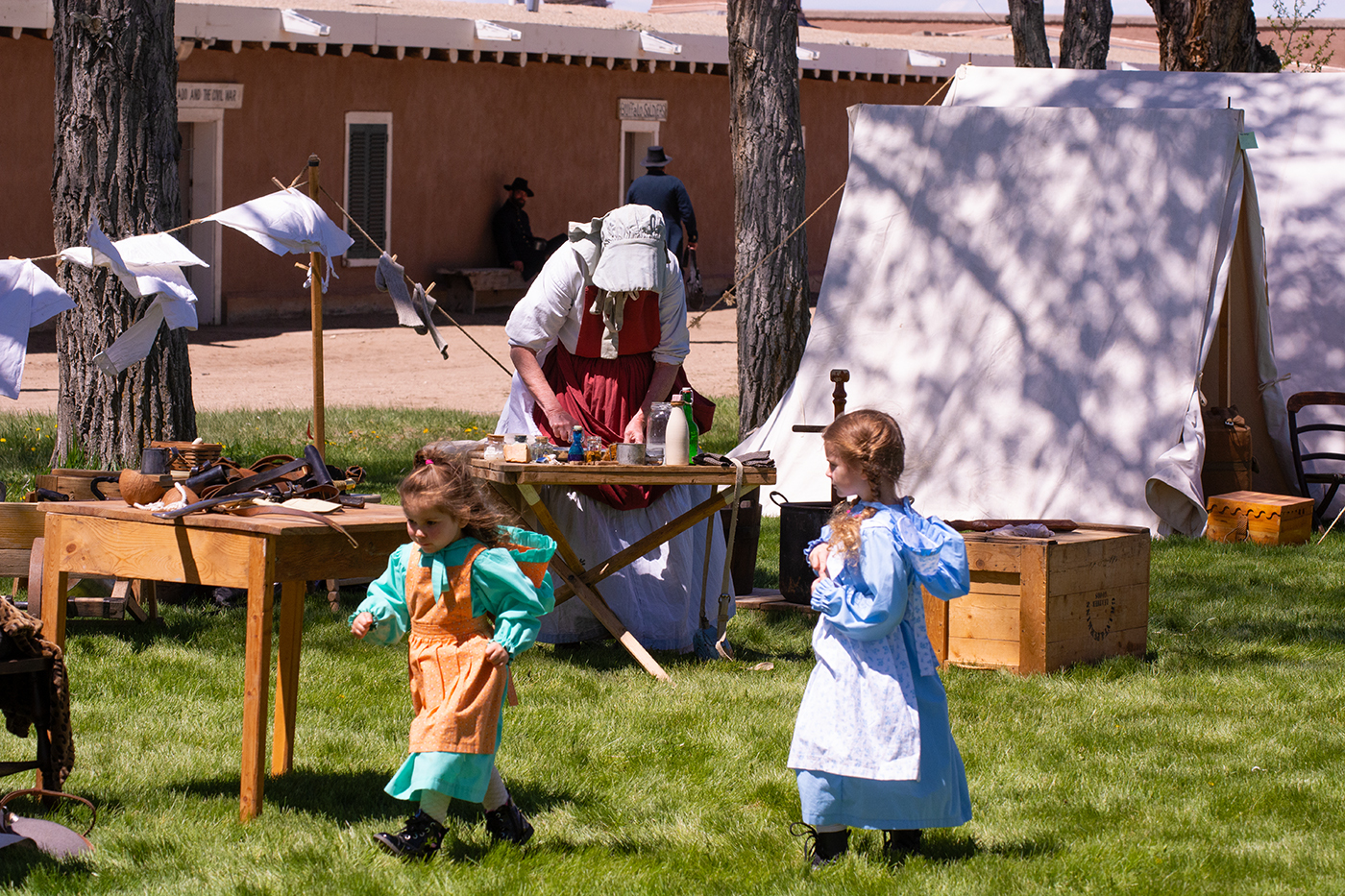 in a reenactment scene at Fort Garland, two little girls walk past wedge tents and a woman doing chores. Historic adobe buildings are in the background. 