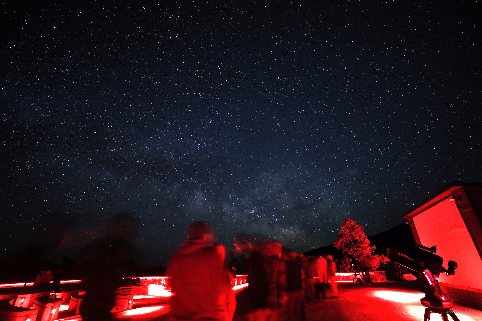 Red lit amphitheater with a colorful Milky Way over visitors waiting in line to view stars.