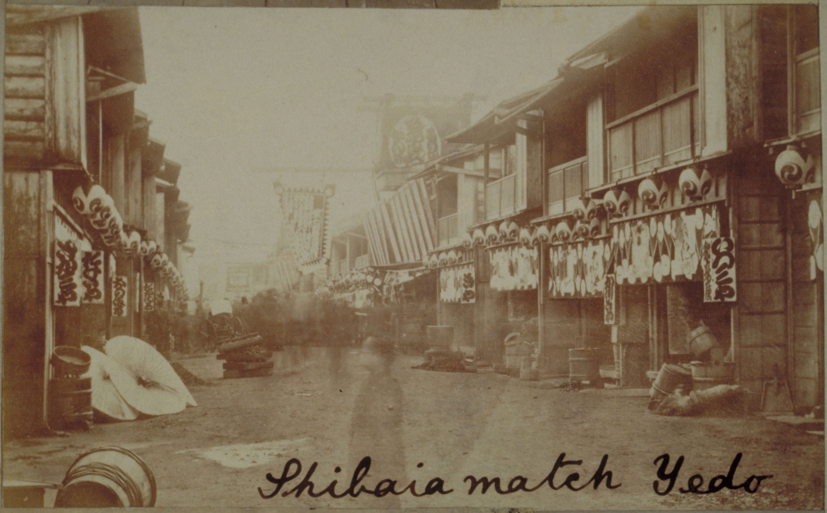 Black-and-white photograph with an inscription representing a town street with wooden buildings decorated by banners and lanterns with Japanese language
