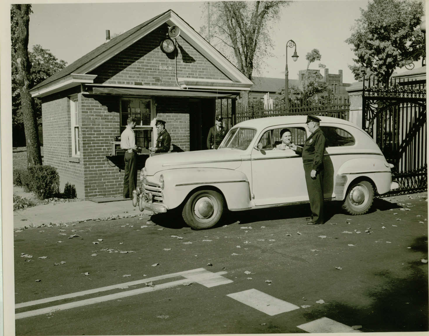 Black and white photo of a car at a gatehouse. The driver and a man in a uniform are exchanging something out the car window. Three men stand by the gatehouse in the background.