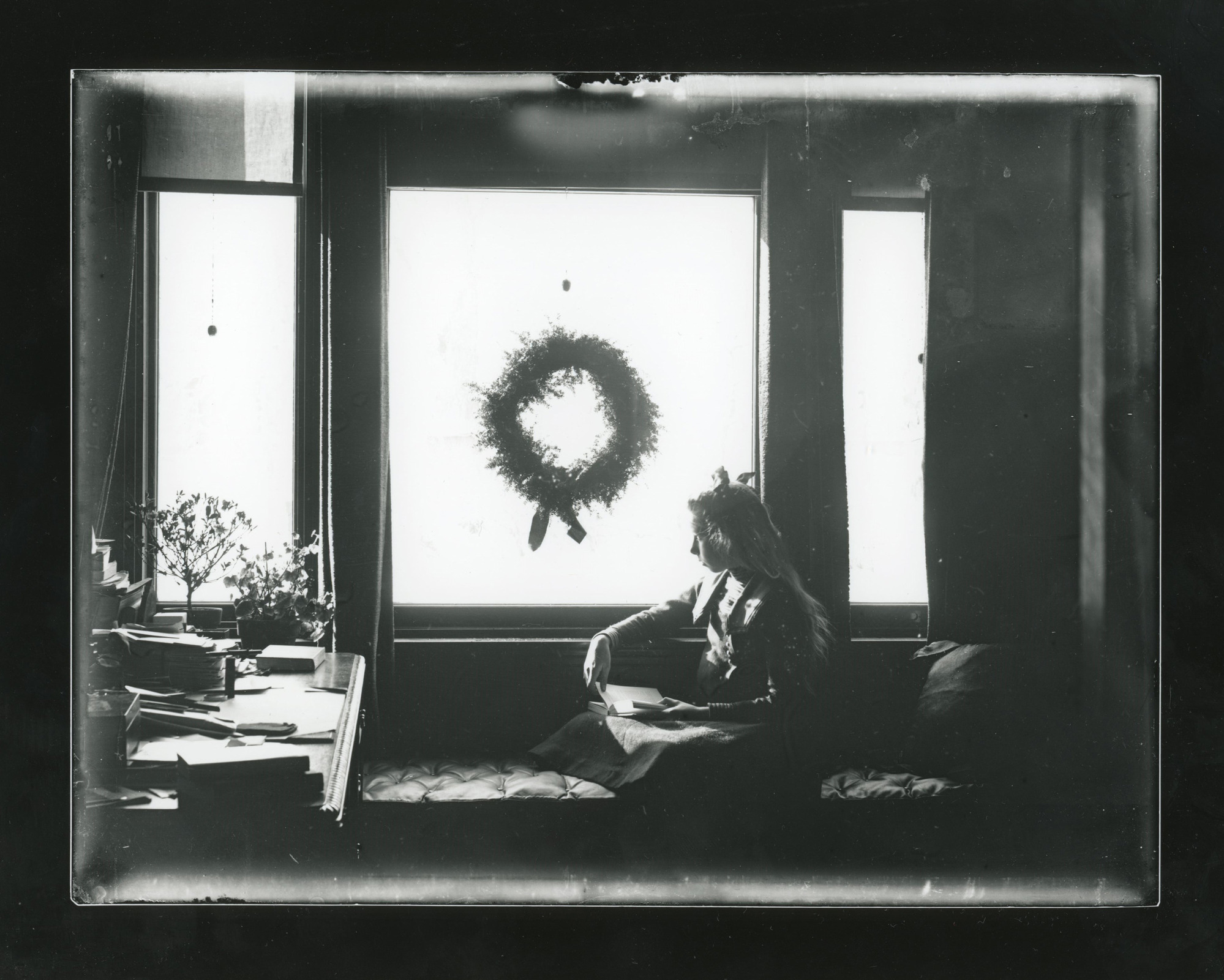 Young white girl sitting on a window seat bench holds a book while looking out a window with a wreath. Desk at left.