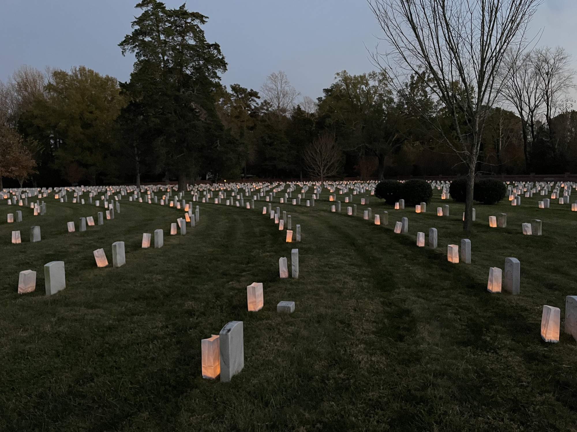 Hundreds of luminaries placed in front of white marble headstones glow in the late evening light.