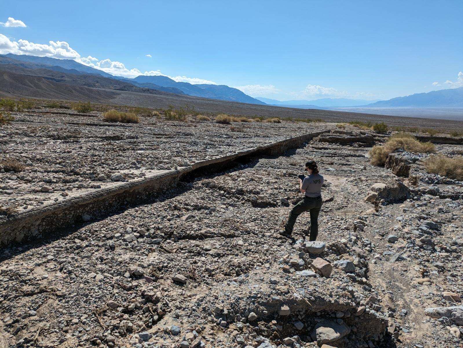 A ranger enters data into an assessment device while standing on the downhill side of a paved road covered in rock and mud debris in a desert landscape with mountains in the distance.