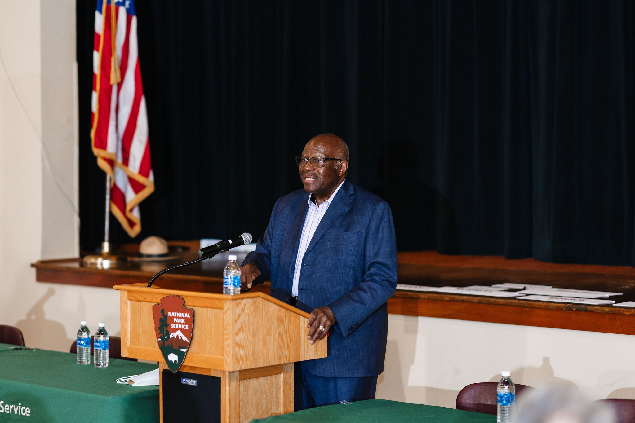 An African American man in a blue suit speaks while standing behind a wooden lectern in front of a wooden stage with the American flag on it