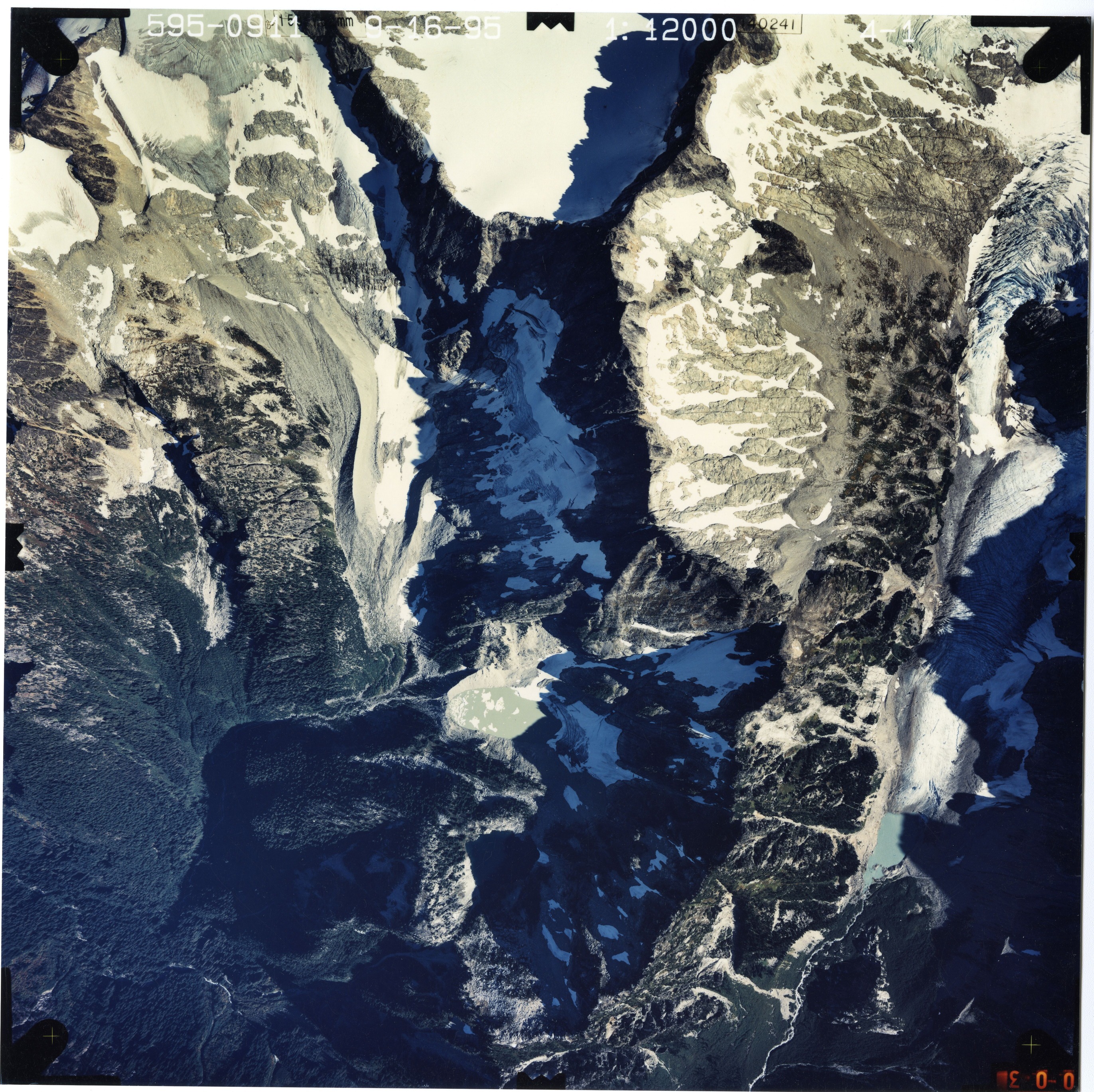 Color aerial photograph of glacier and snowfields on mountain peak, surrounded by forested valleys.