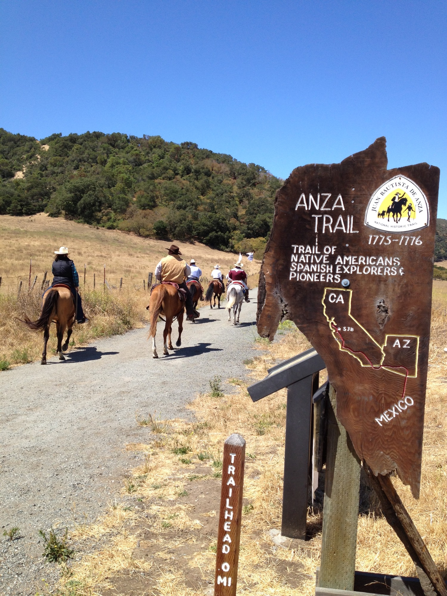 A group of equestrians wearing hats ride down a gravel trail marked with an Anza Trail sign that includes a map of CA and AZ