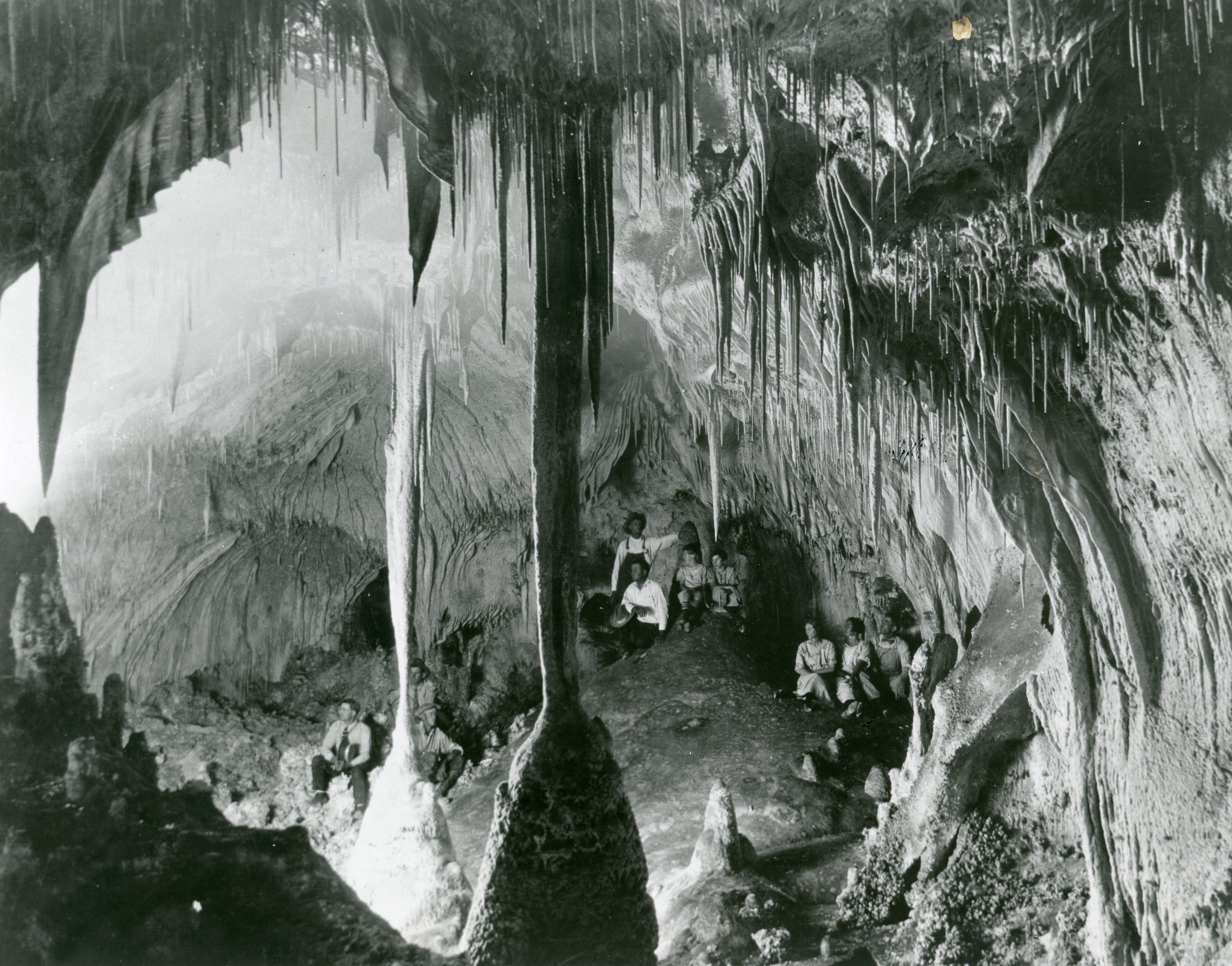 A black and white photograph of early visitors sitting around a cave chamber filled with assorted speleothems.