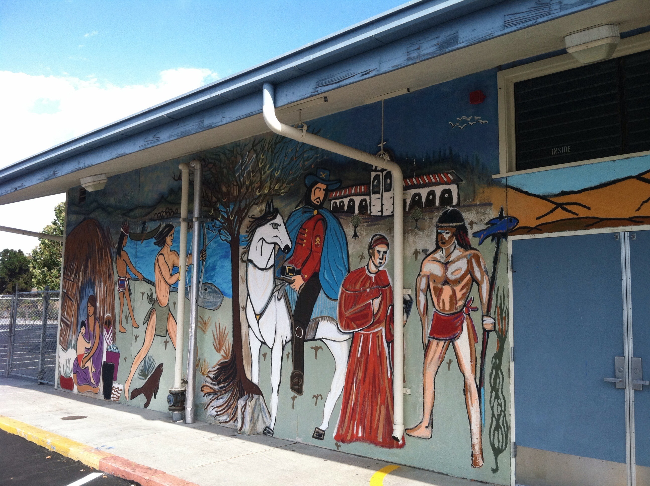 A mural on the side of a building shows indigenous people, a priest, and a Spanish colonizer on a horse