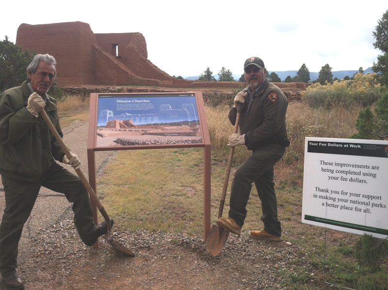 Two men with shovels posing next to an interpretive sign