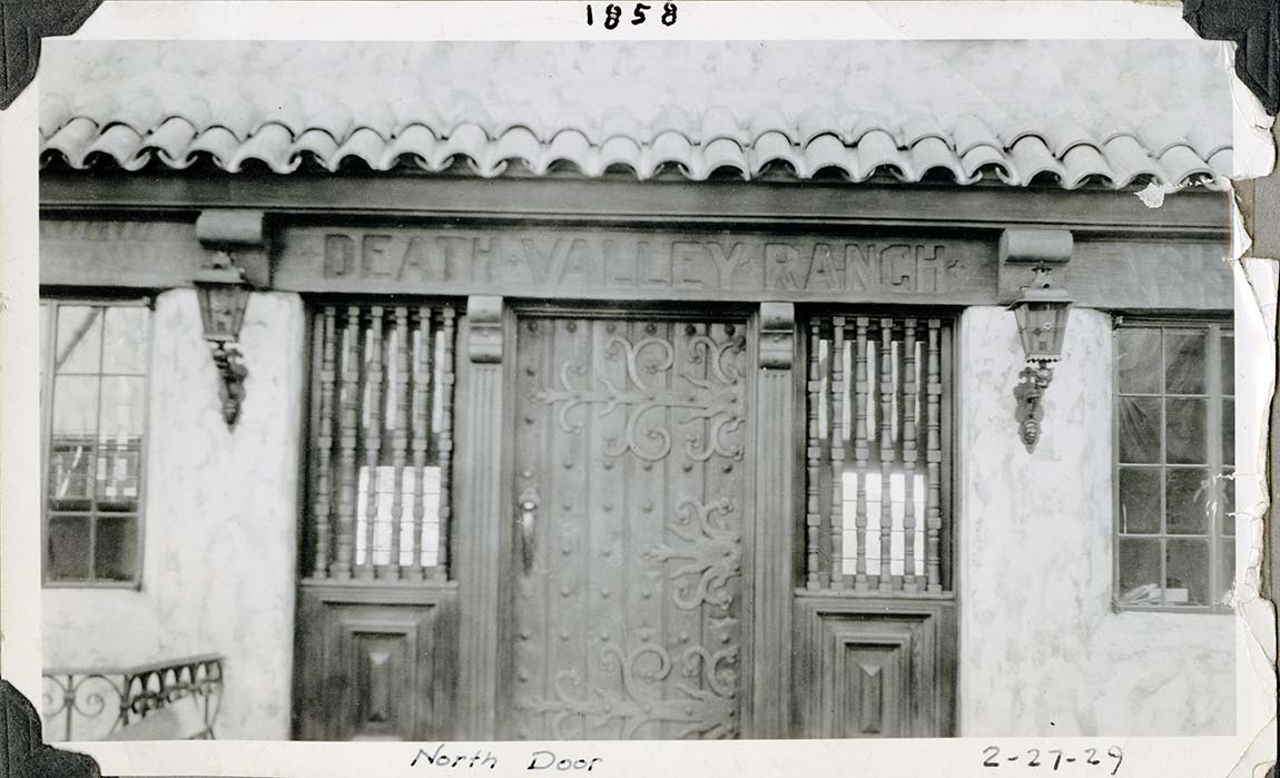 This is an historic black and white photograph from the Scotty's Castle Historic Photograph Collection, Death Valley National Park of door with decorative features.  Spanish tiled eve over door. DEATH VALLEY RANCH carved into header board. Door made of vertical boards secured with hand wrought metal pins and elaborate hinges. Carved wooden vertical balustrades project windows each side door. Wrought iron lantern mounted on bracket each side of door. Inscriptions in black ink along upper and lower borders.