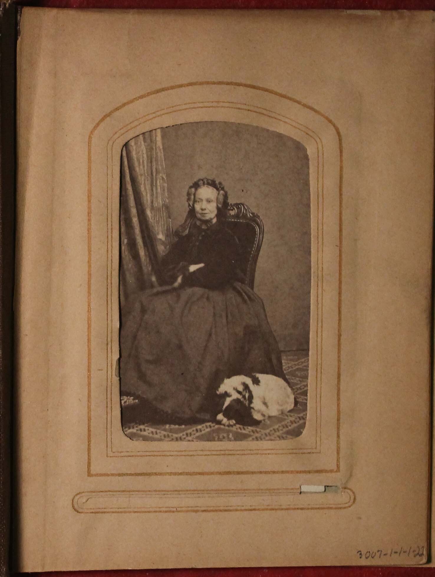 Black and white photograph of older woman sitting posed with small dog curled at her feet.