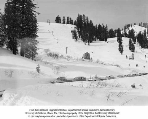 Ski lift with two small structures and several skiers on slope and several parked automobiles on road in foreground.