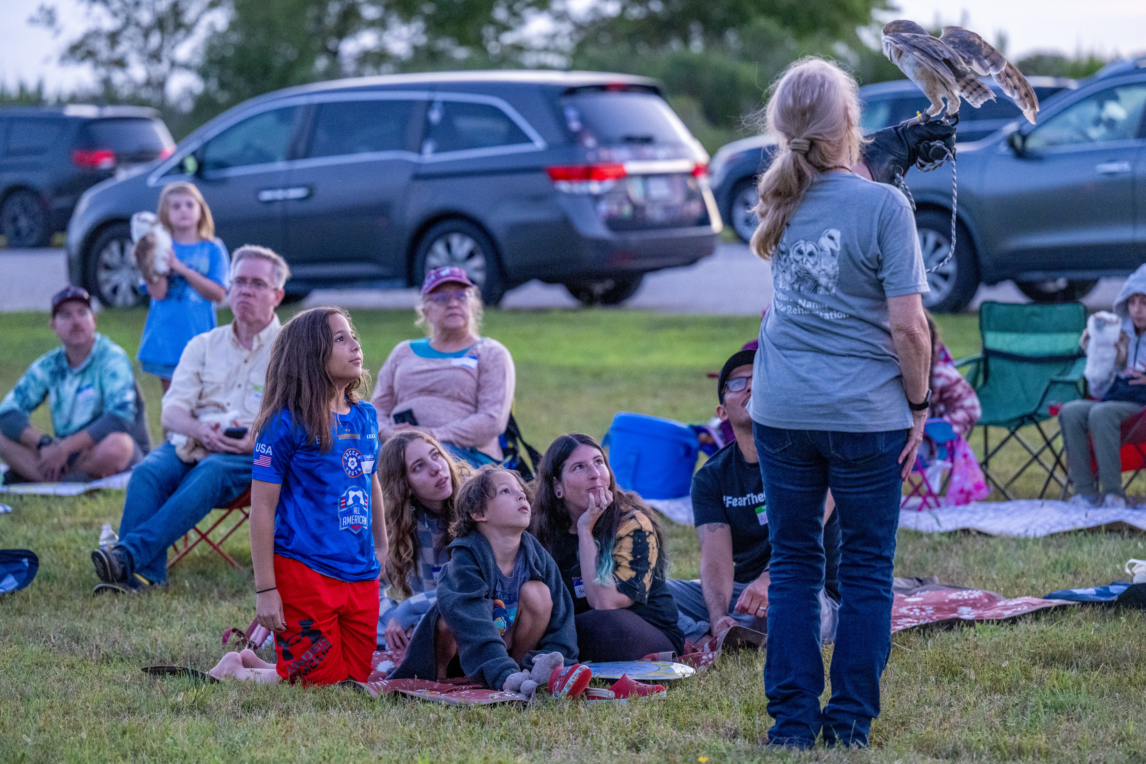 A group of children and adults sitting on a grass watch a woman holding an owl.