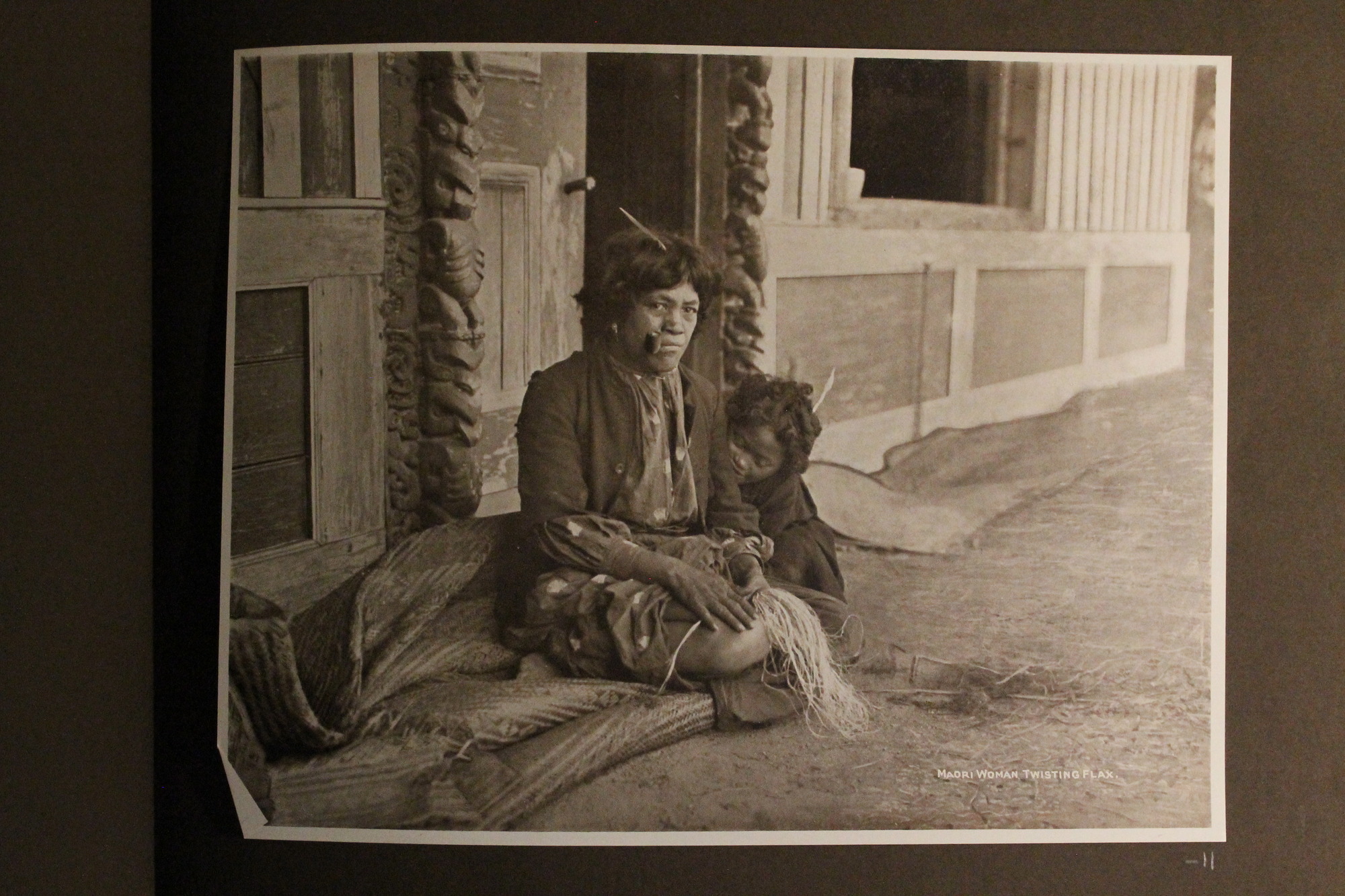 A Maori woman with a child sits near the entrance to a Maori house made of wood.