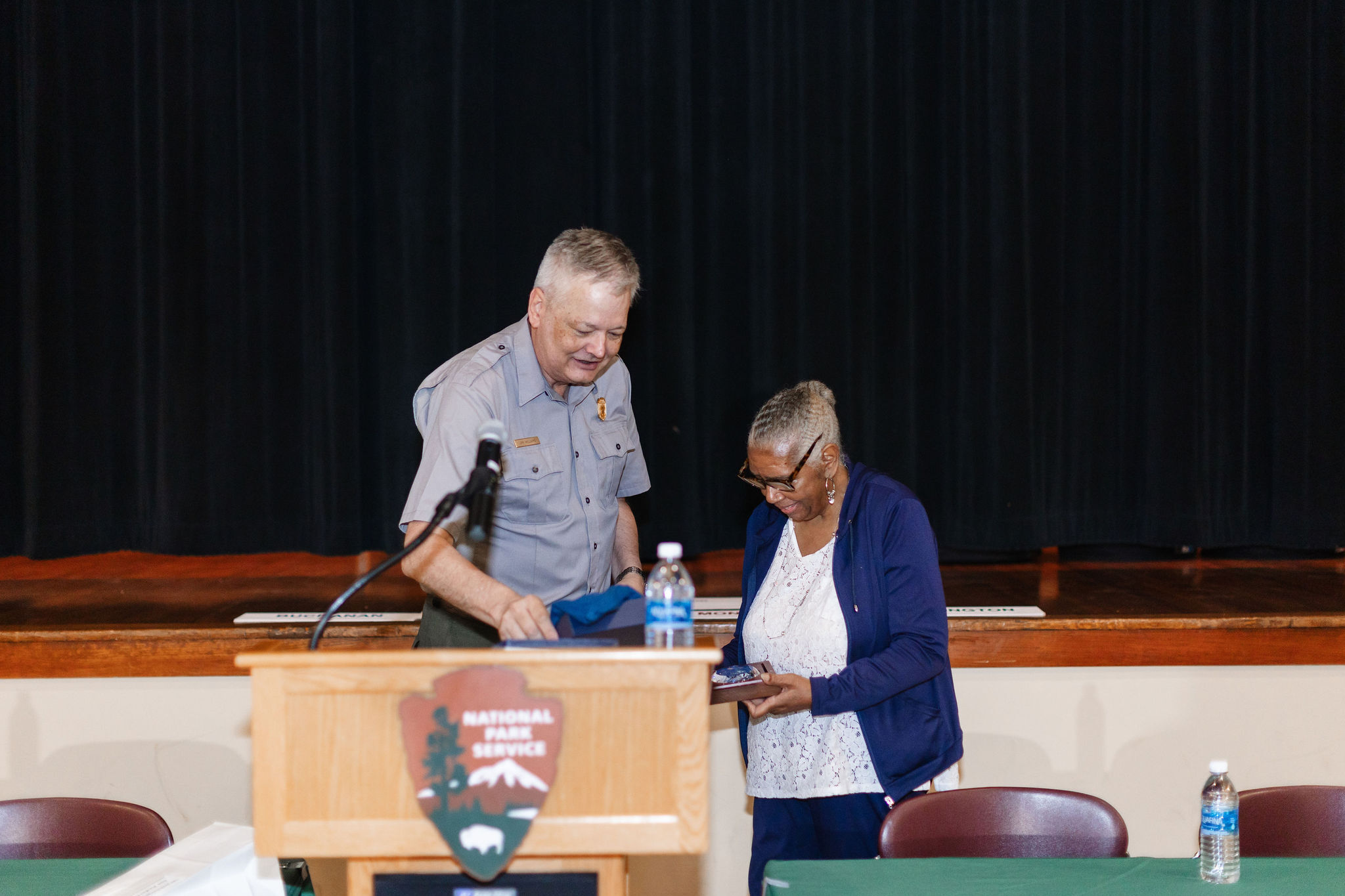 A close-up image of Superintendent Willimas and Marty Paterson behind a podium as they address a crowd that is offscreen. There is a stage behind them with large black curtains that are closed shut.