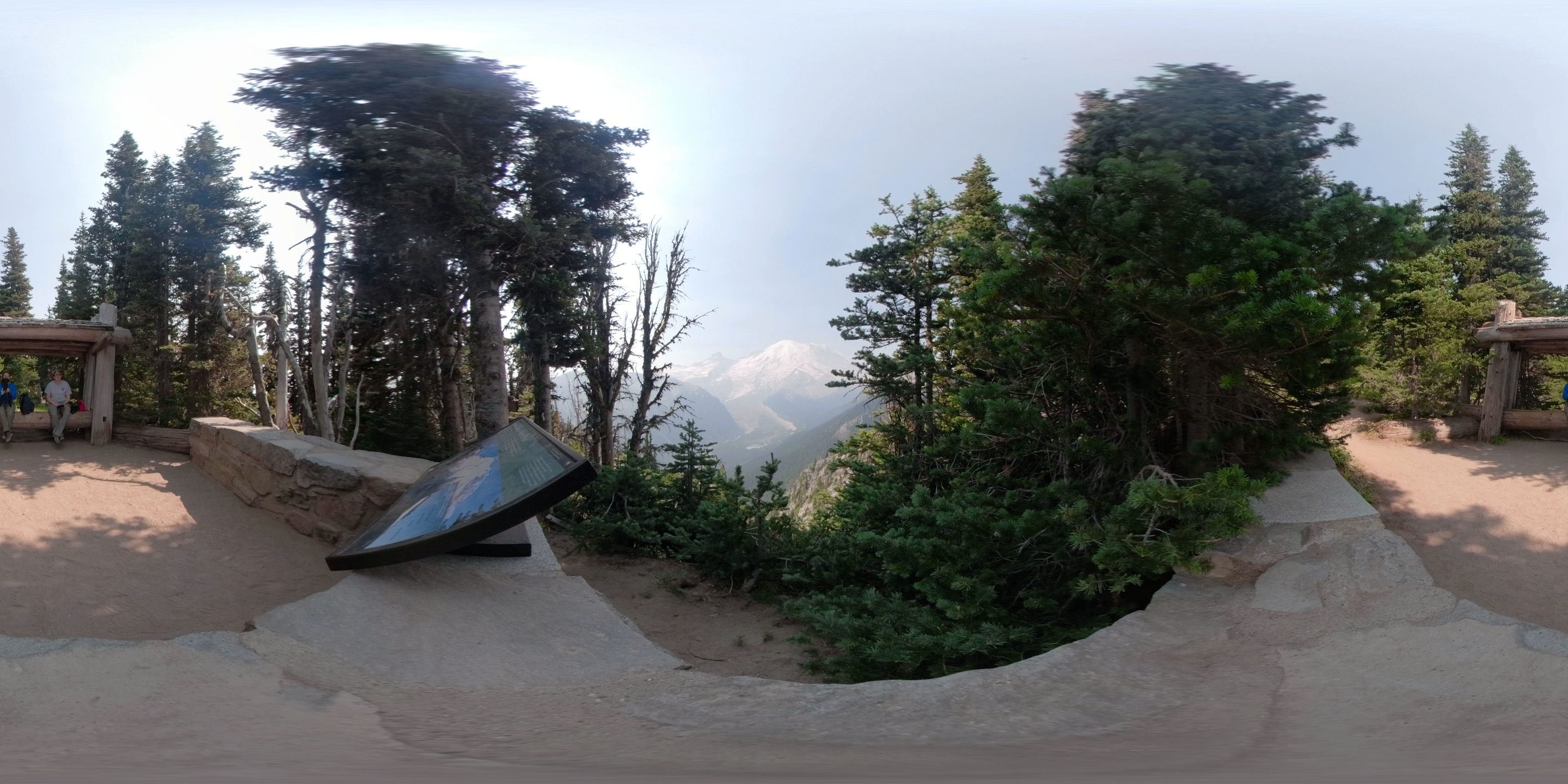 A 360 degree view of Mount Rainier and a glaciated valley from a view point surrounded by a low rock wall with a wayside panel. Two people sit on a large log bench surrounded by trees. 