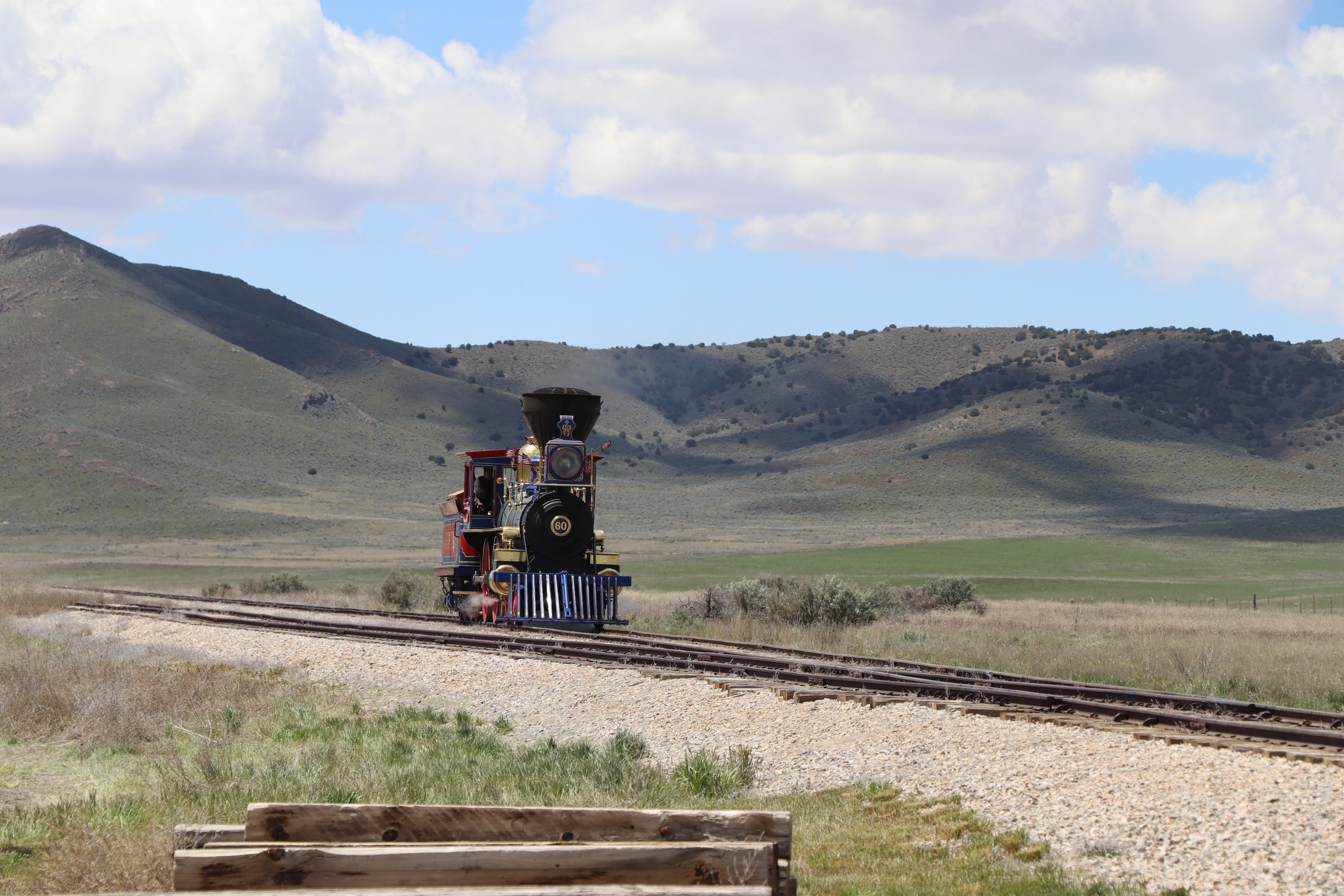 A black, blue, and gold steam locomotive comes down the track. The number "60" is written on the front and white steam billows from around its wheels. Sky, mountain, and sagebrush desert are visible in the background.
