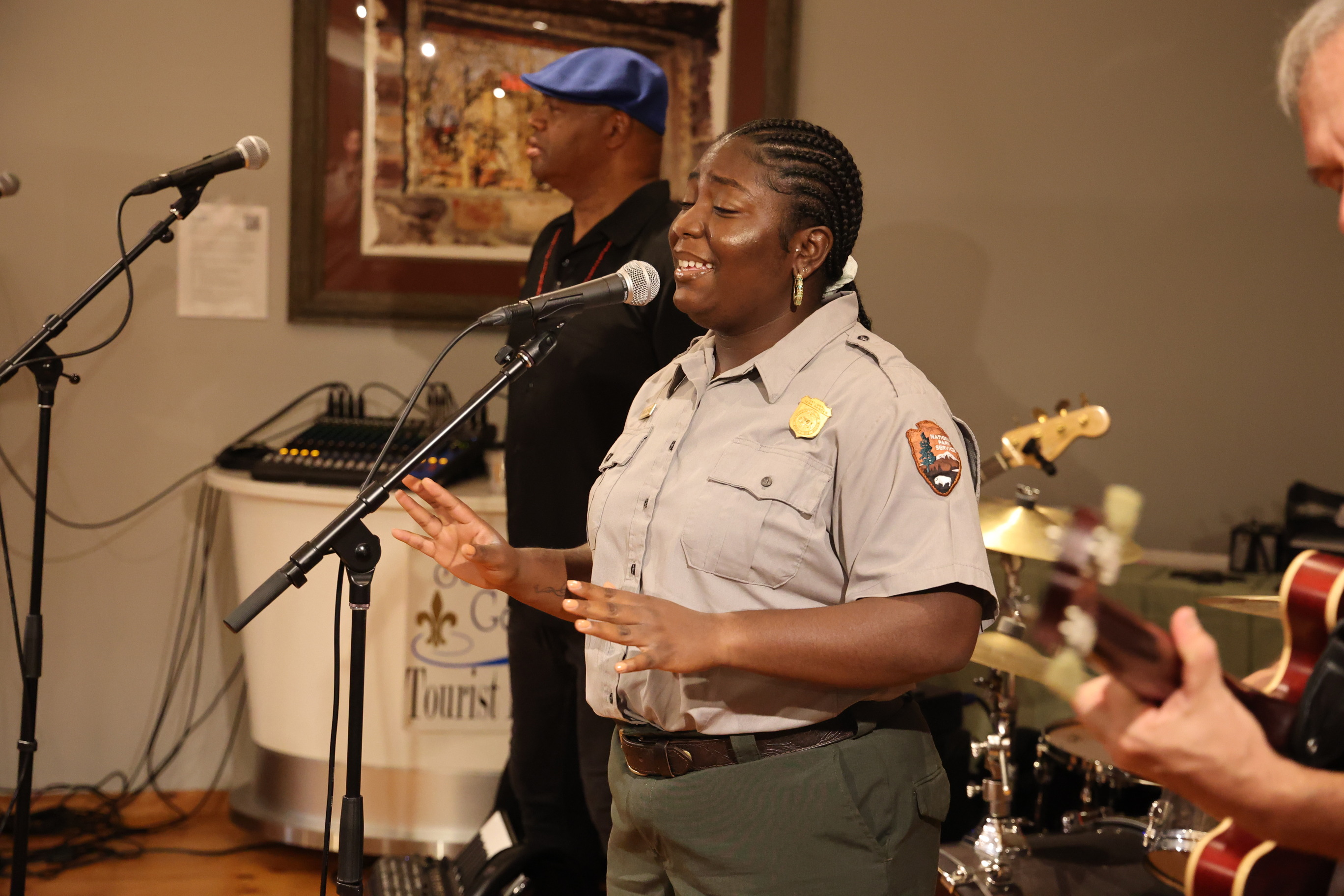 A Park Ranger playing a guitar in the foreground, a Park Ranger singing into a microphone and a person standing back from a microphone. 