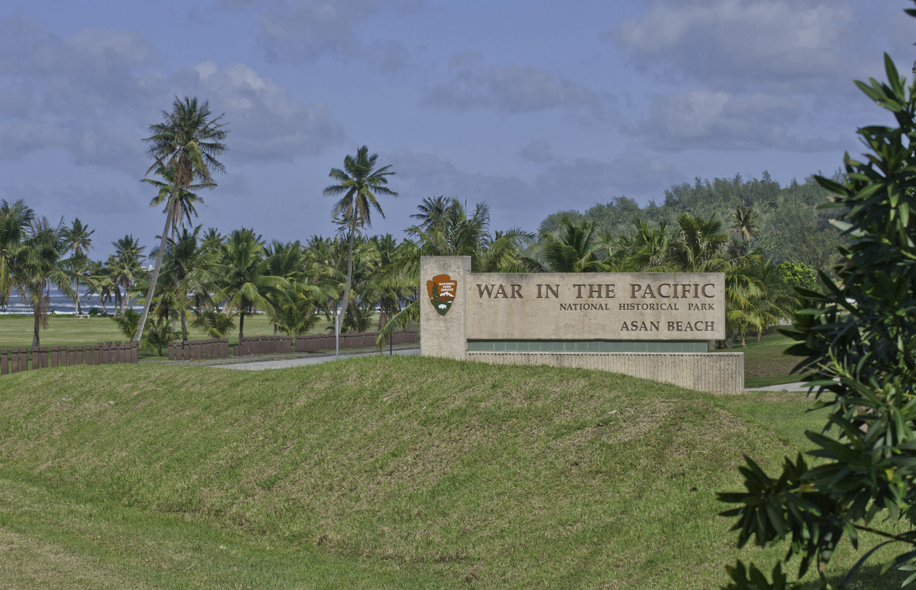 A large concrete sign saying "War in the Pacific National Historical Park Asan Beach" in front of a open green space lined with palm trees.