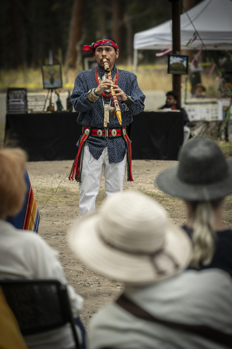 A man in indigenous regalia plays a handmade wooden flute in front of an outdoor audience.