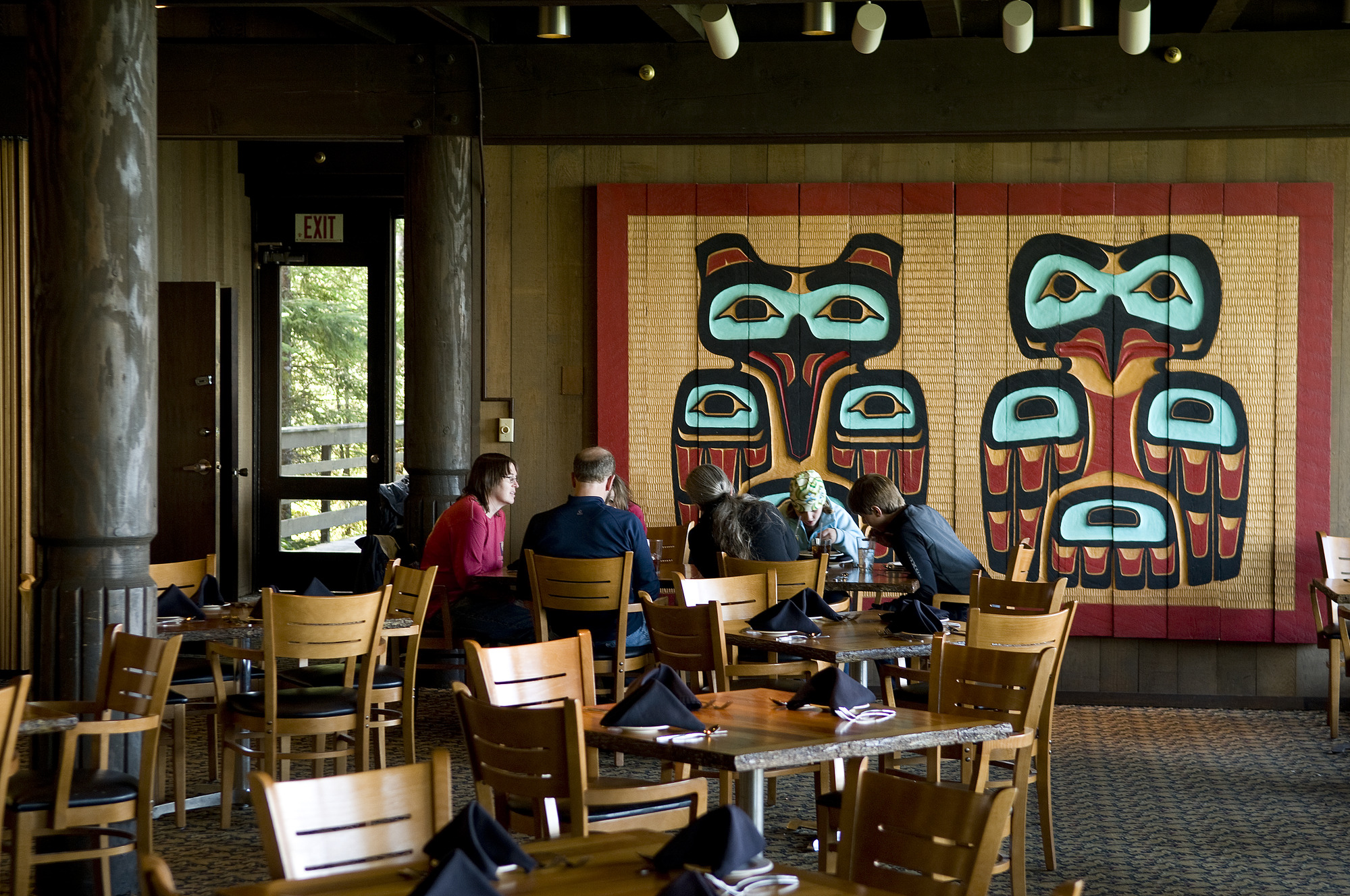 Several people sit at a table in the Glacier Bay Lodge, Tlingit formline art covers the wall behind the group.