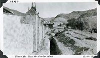 This is an historic black and white photograph from the Scotty's Castle Historic Photograph Collection, Death Valley National Park of excavated trench in foreground parallel to mottled stucco, Spanish style retaining wall. Three men in the ditch, one leaning over outside ditch. Board and bat building with patched tin roof beyond retaining wall. Man on horseback in front of last building. Excavated dirt to right of ditch. Trees on the right. Barren desert mountains in background.  Inscriptions in black ink along upper and lower border.