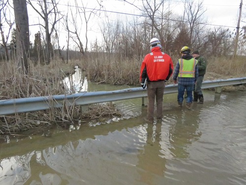 Three men in work clothes stand in ankle-deep floodwater looking over a guard rail. One wears an NPS uniform. Another has a jacket with “U.S. Army Corps of Engineers.” 