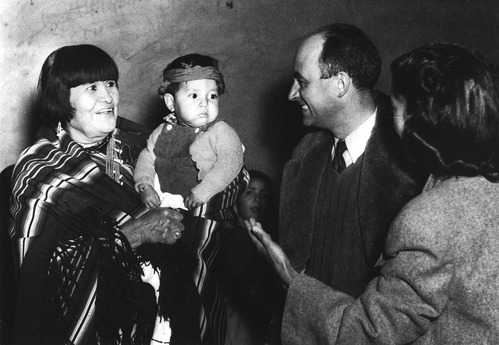Black and white photo of a Pueblo woman standing holding a baby looking towards a man in a suit. A woman with her back to the camera gestures between the two. 