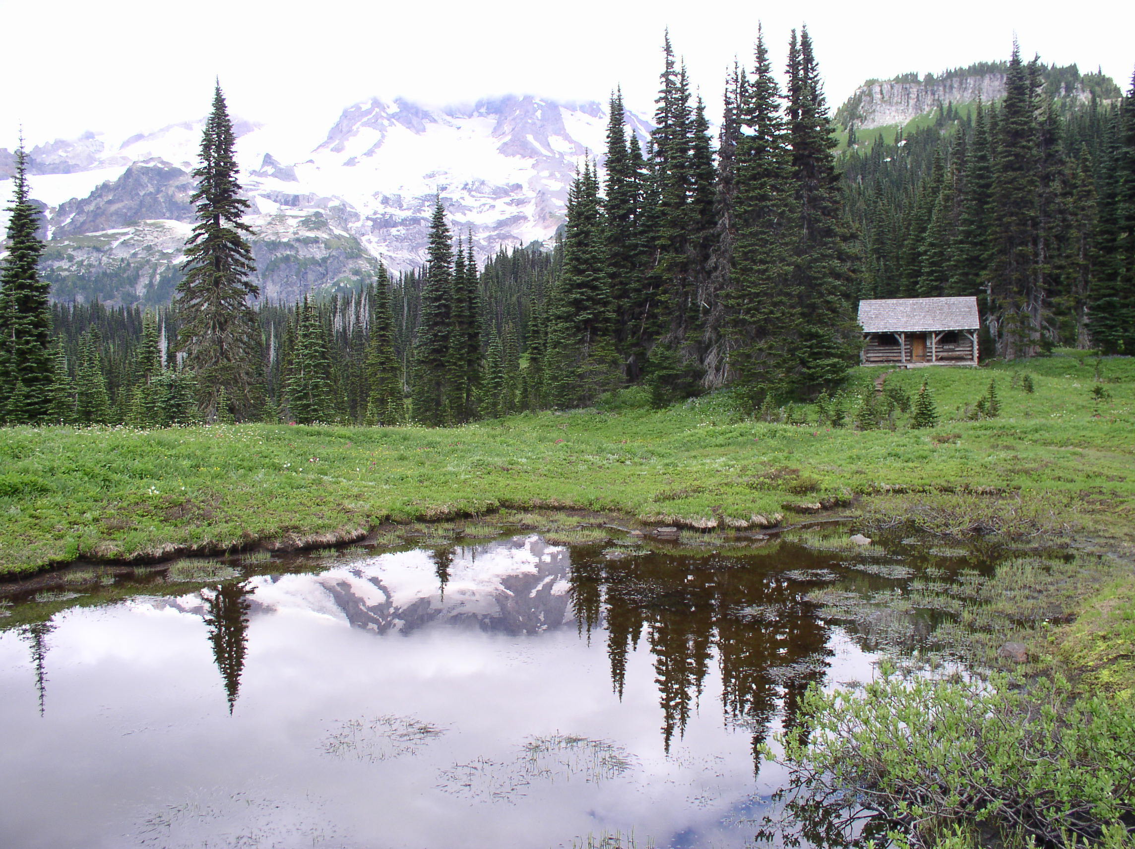 A small log cabin tucked against a line of trees with a lake in front reflecting a mountain partially obscured by clouds. 