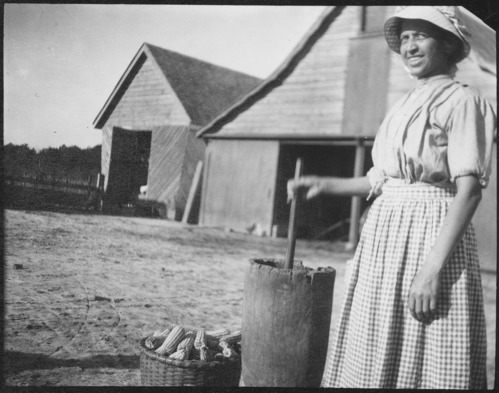 A woman in an apron and sun hat in front of a barn pounding corn in a tall, wooden mortar. 