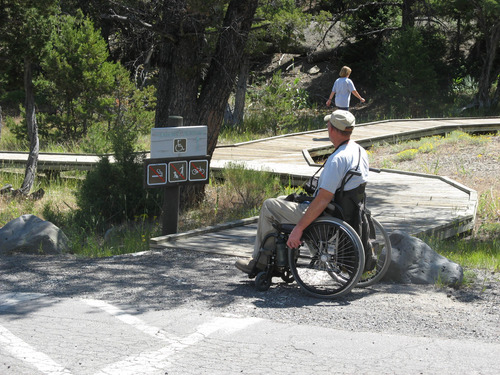 Man in manual wheelchair sits in front of a wooden trail sign in front of a boardwalk.