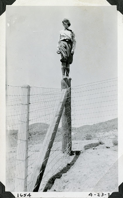 This is an historic black and white photograph from the Scotty's Castle Historic Photograph Collection, Death Valley National Park of Carrie Lee Johnson standing upright on wooden corner fence post, probably at Death Valley Ranch. April 23, 1928. Photographed by Mat Roy Thompson.