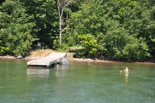 A small wooden dock sticks out into a lake, with a boat tied up. It is surrounded by trees on both sides with a kayaker paddling up to shore. 