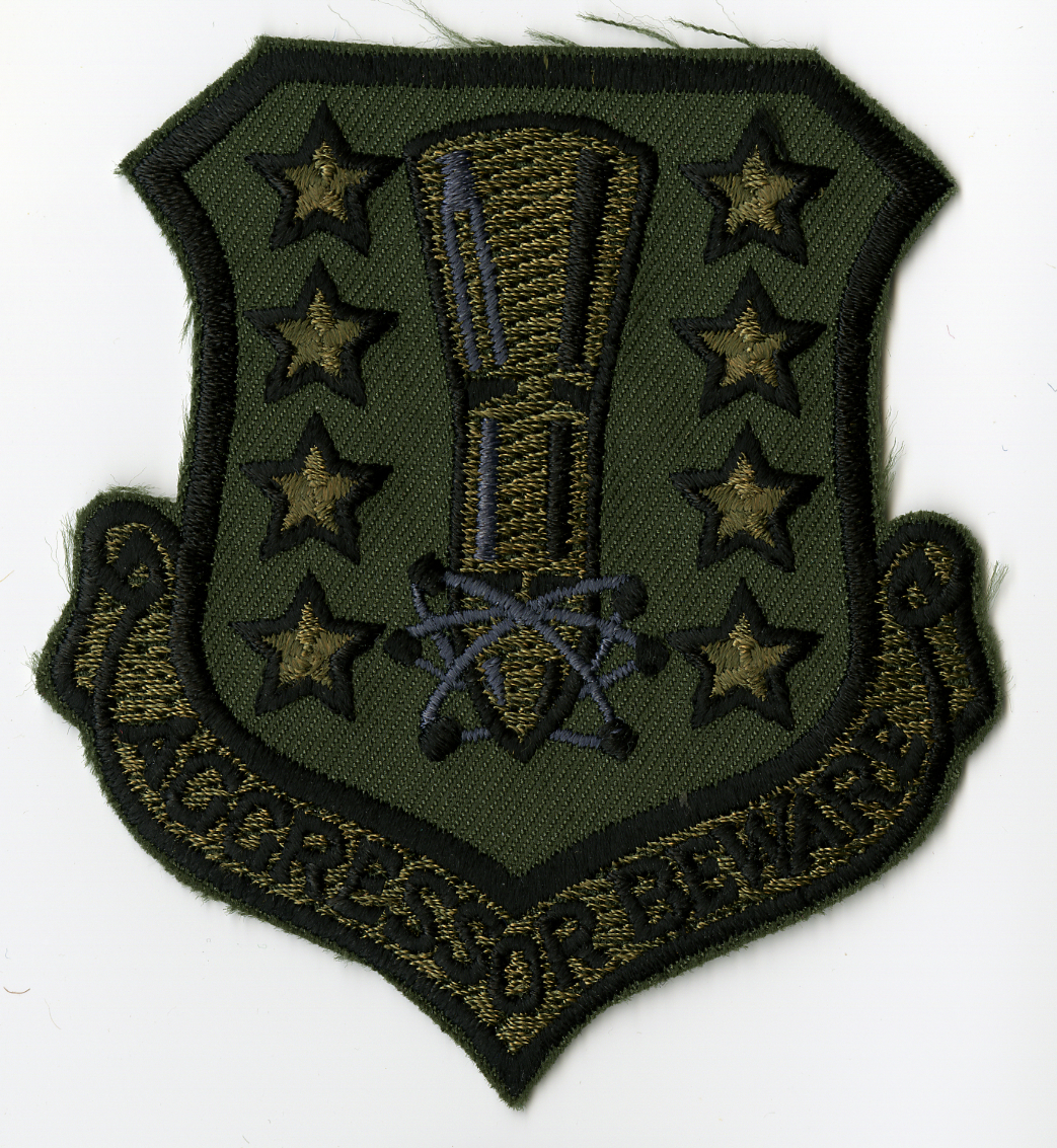 Shield shaped with light blue background and yellow border. Center is yellow, brown, and white nose cone section of missile pointing towards bottom. Nose section has the symbol for atomic science around it. There are four (4) white stars with black edges down each side of the missile. Along bottom is scroll with AGGRESSOR BEWARE" in yellow on white background. Black hook portion of hook & loop fabric on back."

