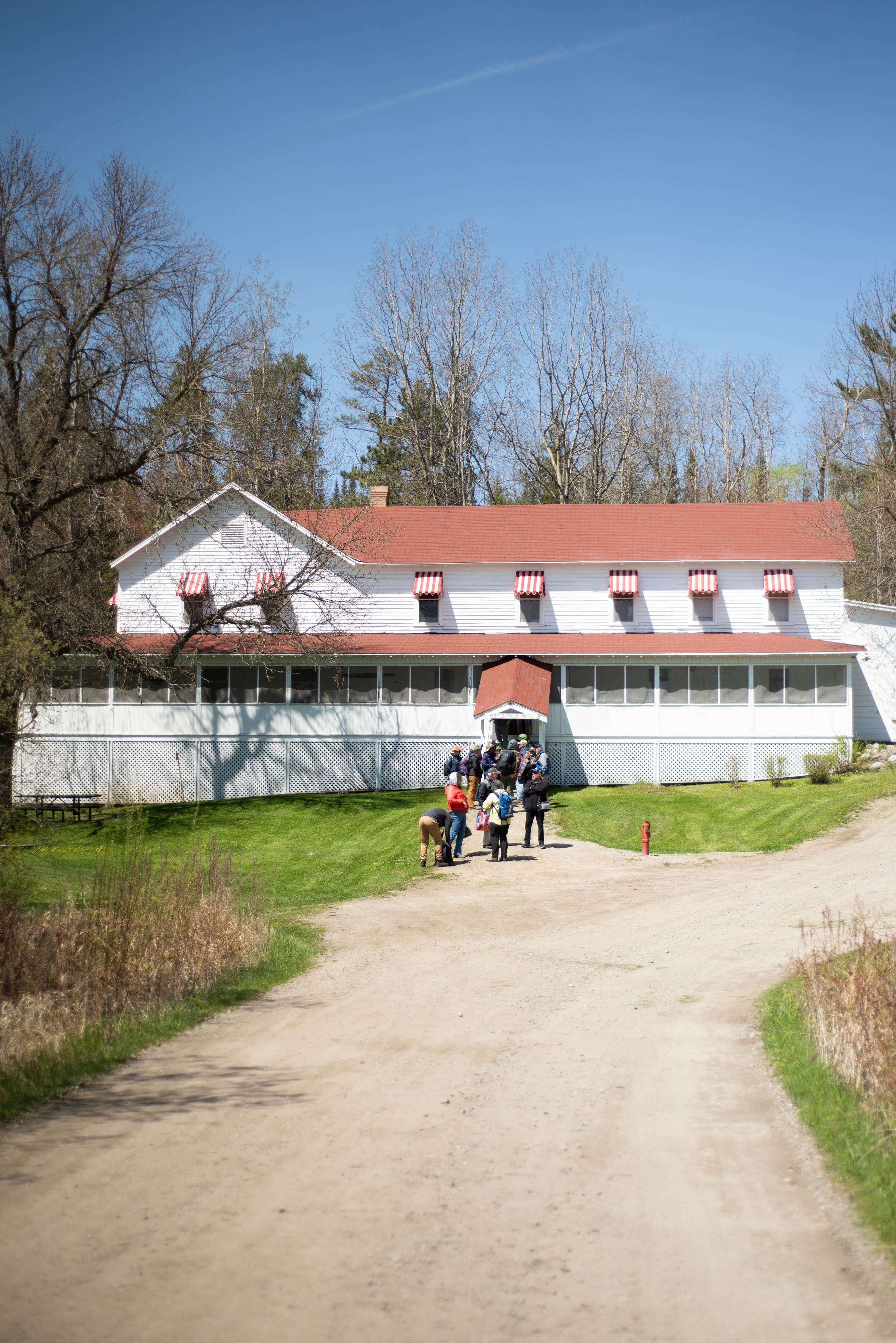 Photograph of a dirt road leading to the front entrance of Ketle Falls Hotel with a red roof, red-and-white striped awnings, and a screened-in porch.